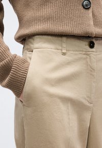 Κοντινό πλάνο ατόμου που φοράει μπεζ παντελόνι και καφέ πλεκτό πουλόβερ, με το ένα χέρι στην τσέπη, εμφανίζοντας τις λεπτομέρειες των κουμπιών στο ρούχο.