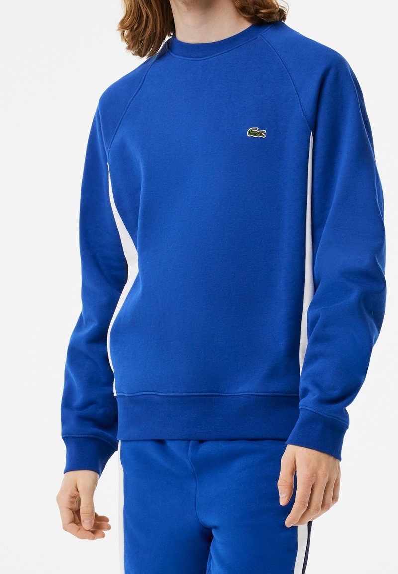 Personne portant un sweat-shirt bleu et un pantalon assorti avec des panneaux latéraux blancs et un petit logo crocodile vert sur la poitrine.