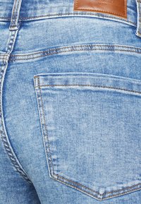 Jeans de ganga de lavagem clara com uma superfície texturizada, apresentando um bolso traseiro e um rótulo de couro castanho na cintura. Costuras em contraste visíveis.