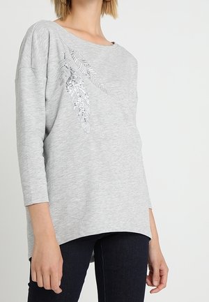 Femme portant un haut gris clair à manches longues avec des motifs de plumes argentées, associé à un pantalon foncé ajusté sur un fond uni.