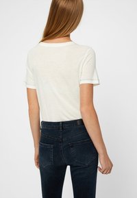 Mujer con cabello largo y lacio, vistiendo una camiseta blanca básica de mangas cortas y unos pantalones vaqueros ajustados de cintura alta de color azul oscuro, vista desde atrás.