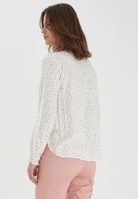 Vit blus med små gula, rosa och gröna blommönster; lättviktsmaterial, långa ärmar med draperade manschetter och rundad nederkant.