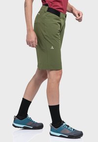 Olivgrüne Shorts mit taillierter Passform, ausgestattet mit einem schwarzen Bund und einem weißen Logo, kombiniert mit blau und schwarzen Schuhen, getragen mit schwarzen Socken.