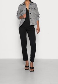 Szara i czarna marynarka w kratę z przednimi kieszeniami, czarny kombinezon i czarne sandały na obcasie. Prosty design, dopasowany krój, gładka faktura.