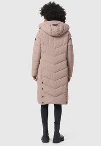 Long padded coat in light pink, featuring a chevron design, hood, and side button closures. Paired with black pants and sturdy boots.