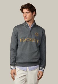 Donkergrijze sweater met half-rits kraag, voorzien van "HACKETT" in gouden letters, gestreepte manchetten en een logo op de borst. Een wit gestreept shirt eronder.