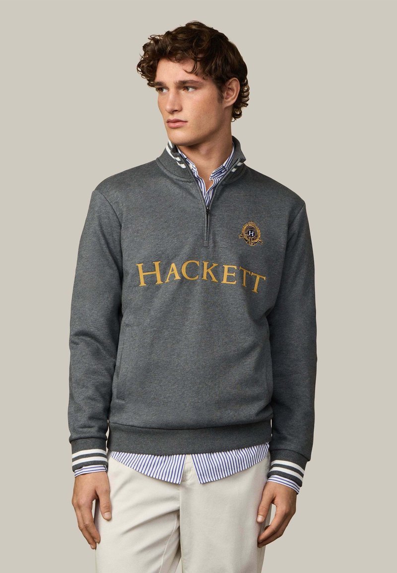 Donkergrijze sweater met half-rits kraag, voorzien van "HACKETT" in gouden letters, gestreepte manchetten en een logo op de borst. Een wit gestreept shirt eronder.