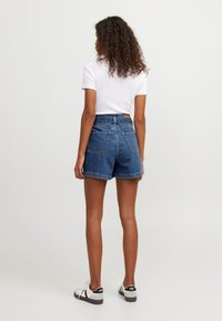 Denim shorts in a medium blue wash, high-waisted with a rolled hem, featuring rear pockets and a subtle stitching detail. Paired with white sneakers.