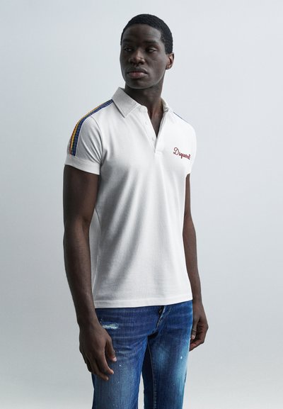 Jeune homme portant un polo blanc avec des rayures arc-en-ciel sur les épaules et un jean bleu, debout devant un fond clair uni.