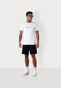 White t-shirt with "ESTEEM MARBELLA" printed in black, paired with black shorts, white sneakers, and sunglasses. Casual, sporty design.