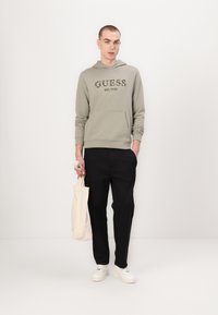 Hellgrüner Kapuzenpullover mit dem Aufdruck "GUESS EST.1981", kombiniert mit schwarzen Hose und weißen Sneakers. Hält eine cremefarbene Tragetasche.