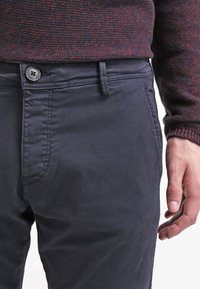 Muž v tmavě modrých kalhotách a tmavě červeném a černém texturovaném svetru, s pravou rukou uvolněně položenou vedle těla.