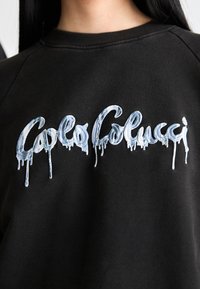 Zwarte hoodie met een druipende, ijzige blauwe geborduurde tekst die "Golo Colucci" leest. De tekst steekt duidelijk af tegen de textuur van de stof.