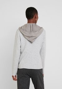 Šedá mikina s šedou kapucí, s hladkou texturou a dlouhými rukávy. Zadní část má přiléhavý střih a viditelné švové detaily.