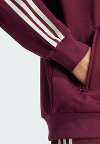 Maroon sweatshirt with white stripes on sleeves, ribbed cuffs, and a front pocket opening. Soft fabric texture, relaxed fit design.
