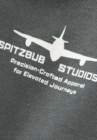 Witte vliegtuigafbeelding en tekst "SPITZBUB STUDIOS Precisie-Gemaakt Kleding voor Verhoogde Reiservaringen" op een grijze gestructureerde stof.