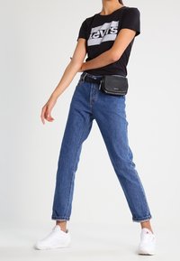 Černé bavlněné tričko s bílým logem "Levi's", vysoké modré džínové kalhoty, černý opasek a bílé tenisky. Černá kabelka přes rameno.