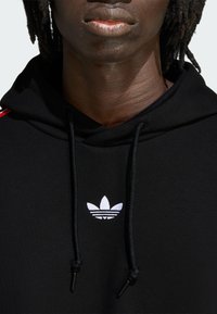 Čierna mikina s kapucňou s bielym vyšívaným logom Adidas na hrudi, vybavená šnúrkami a textúrovanou látkou. Červené akcentové pruhy na rukávoch.
