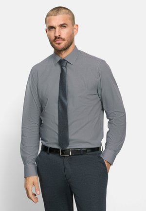Uomo che indossa camicia a maniche lunghe fantasia, cravatta scura e pantaloni scuri con cintura, in piedi con una mano in tasca su sfondo chiaro.