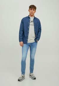 Camisa de mezclilla superpuesta sobre una camiseta gráfica gris claro, combinada con jeans azul claro y zapatillas gris con acentos negros. Elementos de diseño simples.