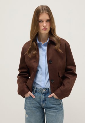 Joven con cabello largo que lleva una chaqueta marrón abotonada, camisa azul claro y jeans desgastados y rotos, de pie con las manos en los bolsillos.