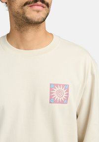 T-shirt a maniche lunghe colore crema con logo quadrato sul petto sinistro, decorato con un sole rosa e scritte blu. Texture in cotone morbido.