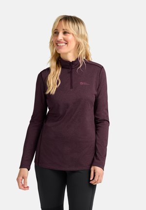 Lächelnde Frau mit langen blonden Haaren, die ein langärmliges, weinfarbenes Zip-Up-Oberteil und schwarze Hosen trägt, steht vor einem schlichten Hintergrund.