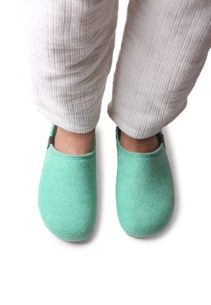 Green felt slippers with a smooth texture, rounded shape, and stitching detail, paired with white, textured pants.