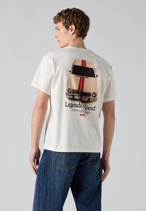 Joven con camiseta blanca con coche vintage y texto "Legenda Speed" en la espalda y vaqueros azules, de pie frente a un fondo liso.
