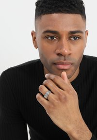 Stříbrný prsten s vyrytým designem, na pravé ruce. Texturovaný černý svetr s proužky. Model má krátké kudrnaté vlasy a neutrální výraz.