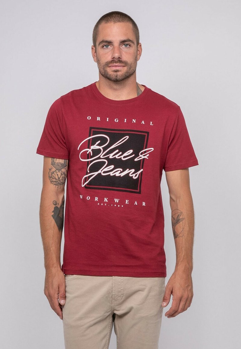 T-shirt en coton rouge avec un imprimé graphique comportant le texte "Blue & Jeans" et "Original Workwear" en blanc et noir, col rond, manches courtes.