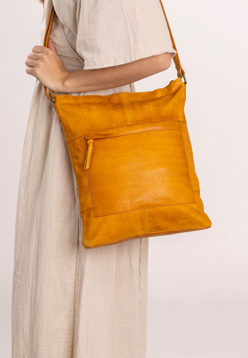 Bolso de hombro de cuero amarillo con una textura suave, que cuenta con un bolsillo frontal con cremallera y una forma plana, llevado a través de una correa ajustable.
