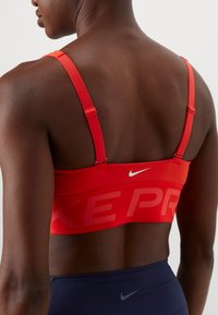 Red sports bra with adjustable straps, featuring a large "NIKE PRO" logo in a tonal pattern on the band, made from smooth, stretchy material.