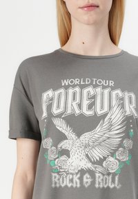 T-shirt gris à manches courtes, avec un col rond, et un design graphique représentant un aigle, des roses, et le texte "FOREVER ROCK & ROLL."