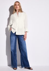 Weißes Leinenhemd mit langen Ärmeln und Knöpfen, kombiniert mit weit geschnittenen blauen Denim-Jeans und schwarzen Rutschen. Glatter, luftiger Stoff.