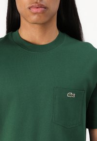 T-shirt verde in cotone con scollo a girocollo, dotato di una piccola tasca sulla sinistra del petto e un logo del coccodrillo ricamato in bianco e rosso.