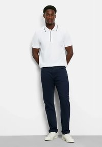 Weißes Polohemd mit schwarzen Kragenakzenten, kombiniert mit marineblauen Hosen und weißen Turnschuhen. Das Material wirkt glatt und eng anliegend.