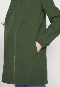 Veste verte à manches longues avec fermeture éclair frontale, poches latérales, poignets élastiques et taille ajustable par cordon, portée avec la main dans la poche.