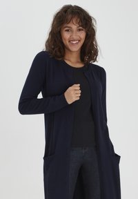 Dlouhý navy cardigan, pletená látka, otevřený přední díl, žebrované okraje, dvě přední kapsy, kombinovaný s černým topem a tmavými džíny.