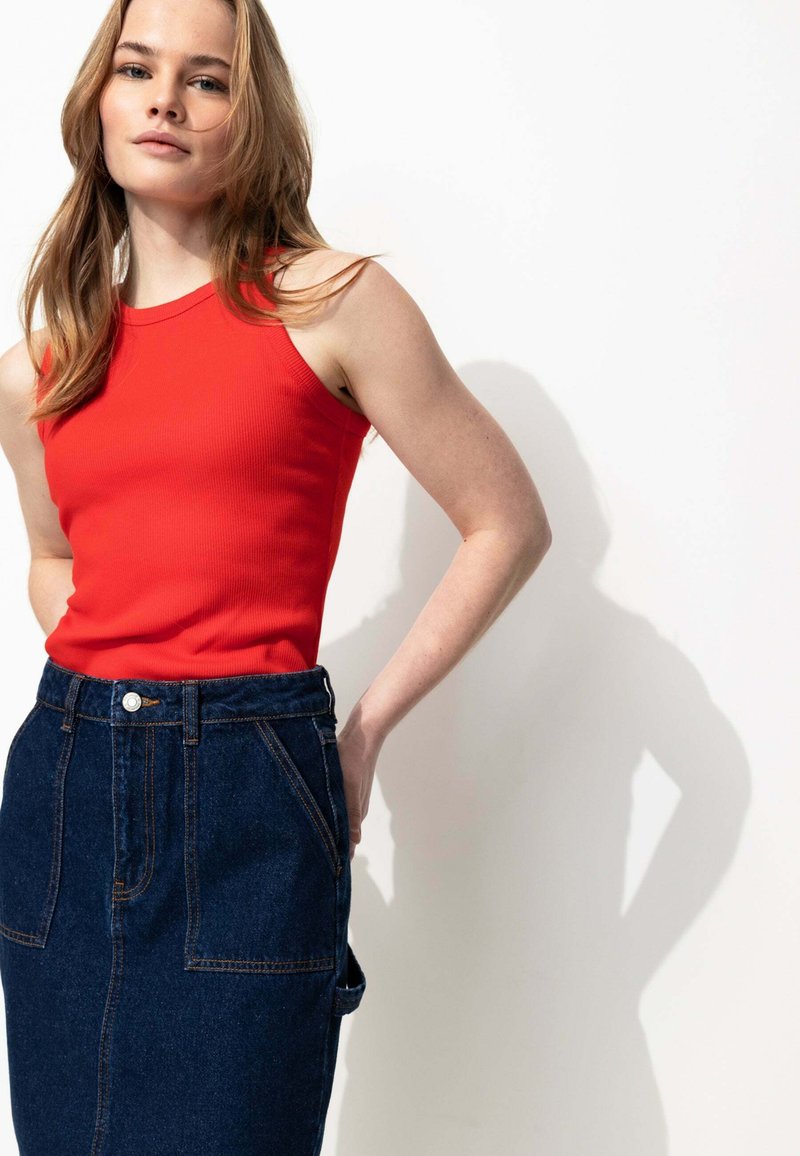 Jeune femme aux longs cheveux portant un haut sans manches rouge et une jupe en jean foncé taille haute, se tenant devant un fond blanc uni.