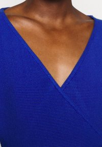 Osoba o ciemnej karnacji, ubrana w głęboki top w kolorze kobaltowym z dekoltem w serek i prążkowanym wzorem, odsłaniająca obojczyk i górną część klatki piersiowej.