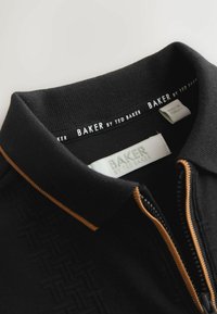 Sort polo-shirt med et tekstureret mønster, kontrasterende guld syninger, lynlås foran og et mærket krave-tag.