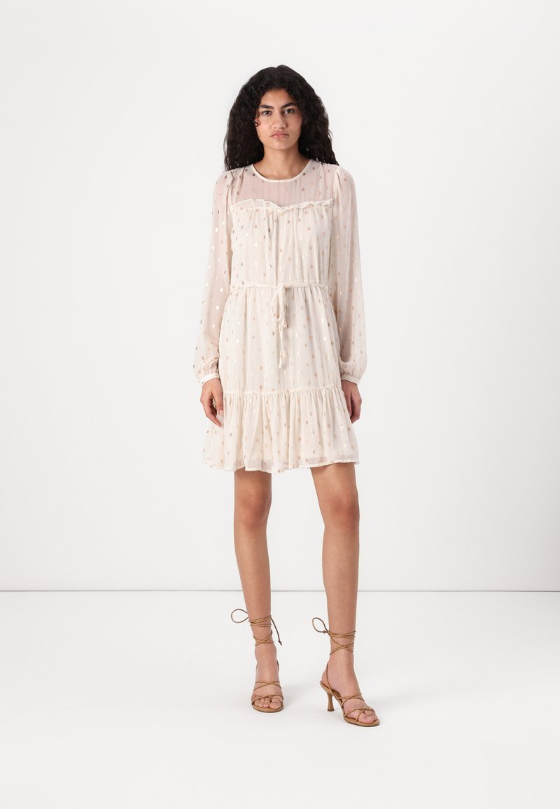Robe crème à pois, à manches longues, avec un col à volants, une taille froncée et une jupe à volants. Portez-la avec des escarpins beiges à brides.