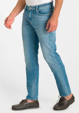 Mann trägt hellblaue Slim-Fit-Jeans und braune Leder-Bootsschuhe, steht mit einem Fuß leicht nach vorne.