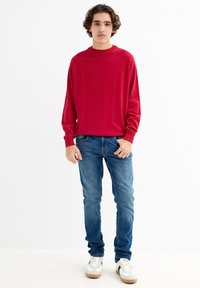 Suéter rojo de canalé con cuello redondo, combinado con jeans azules y zapatillas blancas con detalles en negro. La tela parece suave y texturizada.