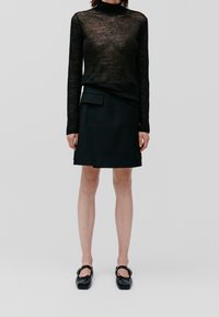 Schwarzes Langarmoberteil aus transparentem, strukturiertem Stoff, kombiniert mit einem schwarzen, hoch taillierten Rock mit seitlicher Taschenapplikation und flachen Schuhen.