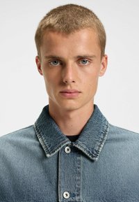 Ljusblå denimjacka med knapprad fram, spetsig krage och två silverknappar. Har en texturerad tyg med synliga sömmar.