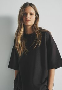 Pārmērīgs melns kokvilnas T-krekls ar plašām piedurknēm un apaļu dekoltē. Mīksta tekstūra, ikdienišķs dizains un brīvs piegriezums.