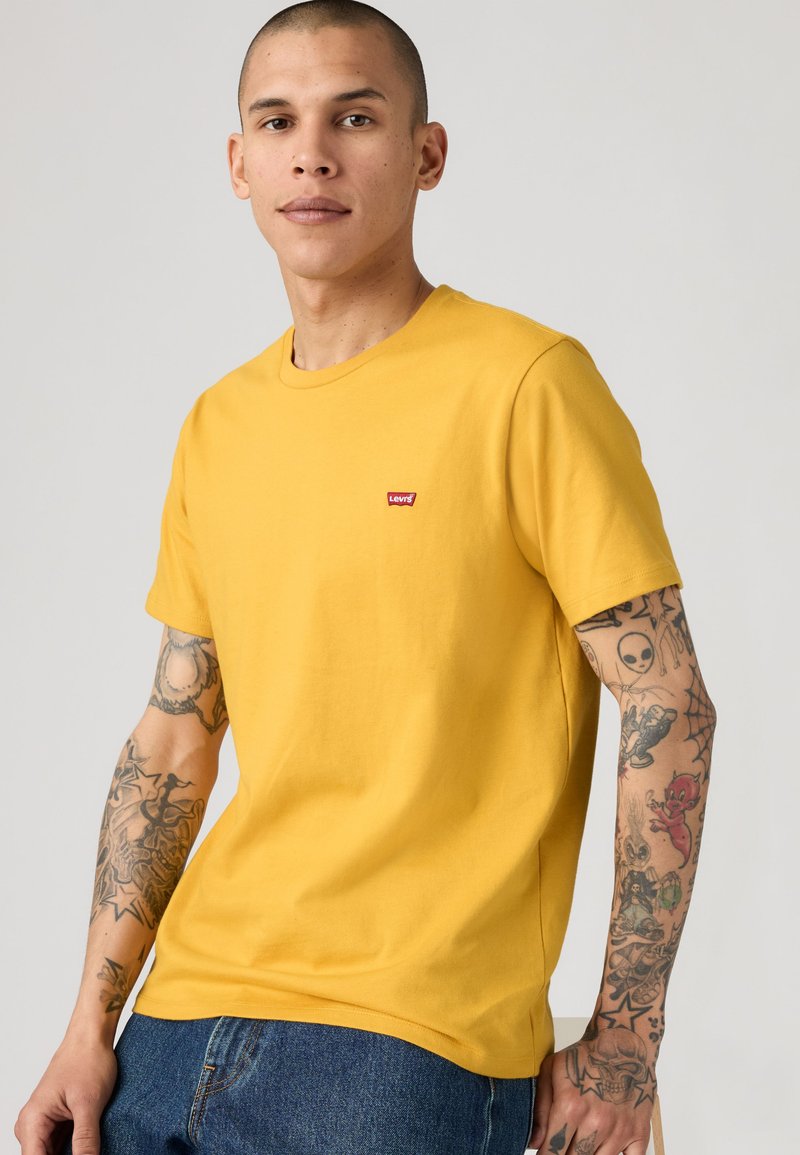 Homme avec coupe buzz, portant un t-shirt jaune Levi's et un jean bleu, montrant ses bras tatoués sur un fond clair uni.