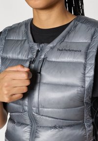 Pamušaluota tamsiai pilka liemenė su užtrauktuku priekyje, pasižyminti siuvinėtu raštu ir „Peak Performance“ logotipu, užsiūtu kairiajame viršutiniame kampe.